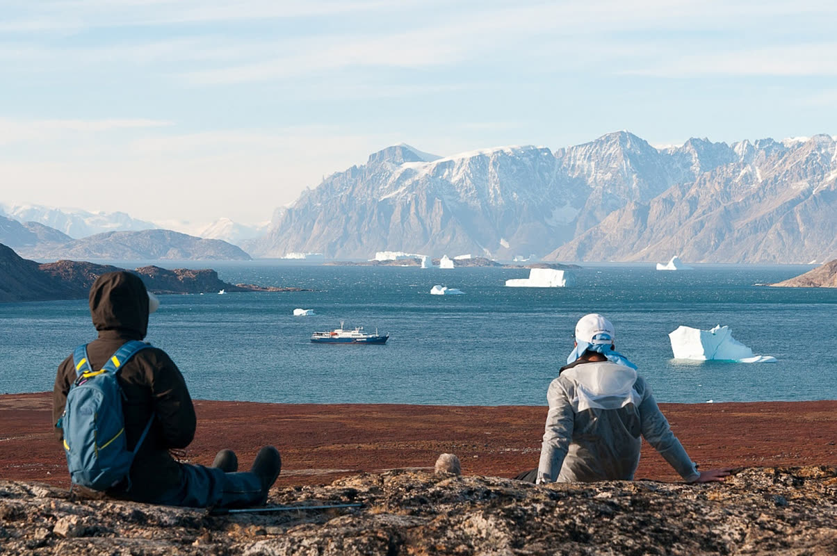 Zwei Gäste sitzen an einem Hang über dem Fjord bei Sydkap, im Wasser das Expeditionsschiff und Eisberge